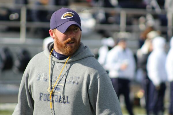 A focused football coach in a hoodie and cap on the sidelines of a stadium, ready to lead the team.